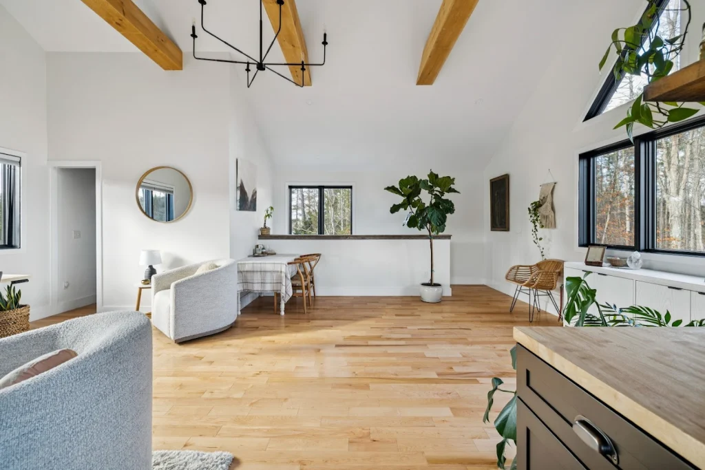 Spacious open-concept living area with wood beams, light wood floors, and abundant natural light from large windows