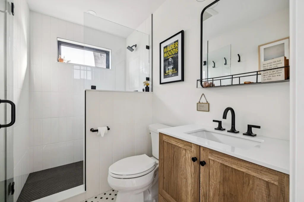 Modern white bathroom with wood vanity, black fixtures, shower, toilet, and framed art