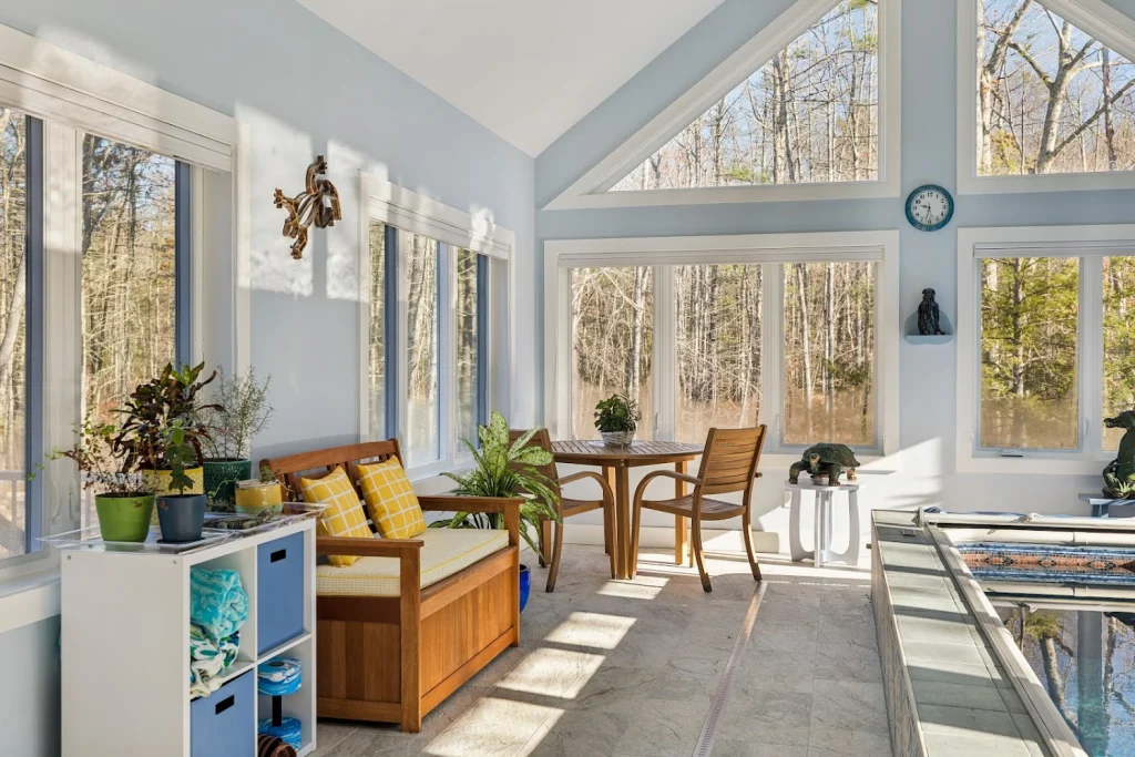 Sun-drenched room with tall windows overlooking a forest, featuring an indoor swim spa, dining set, and plants
