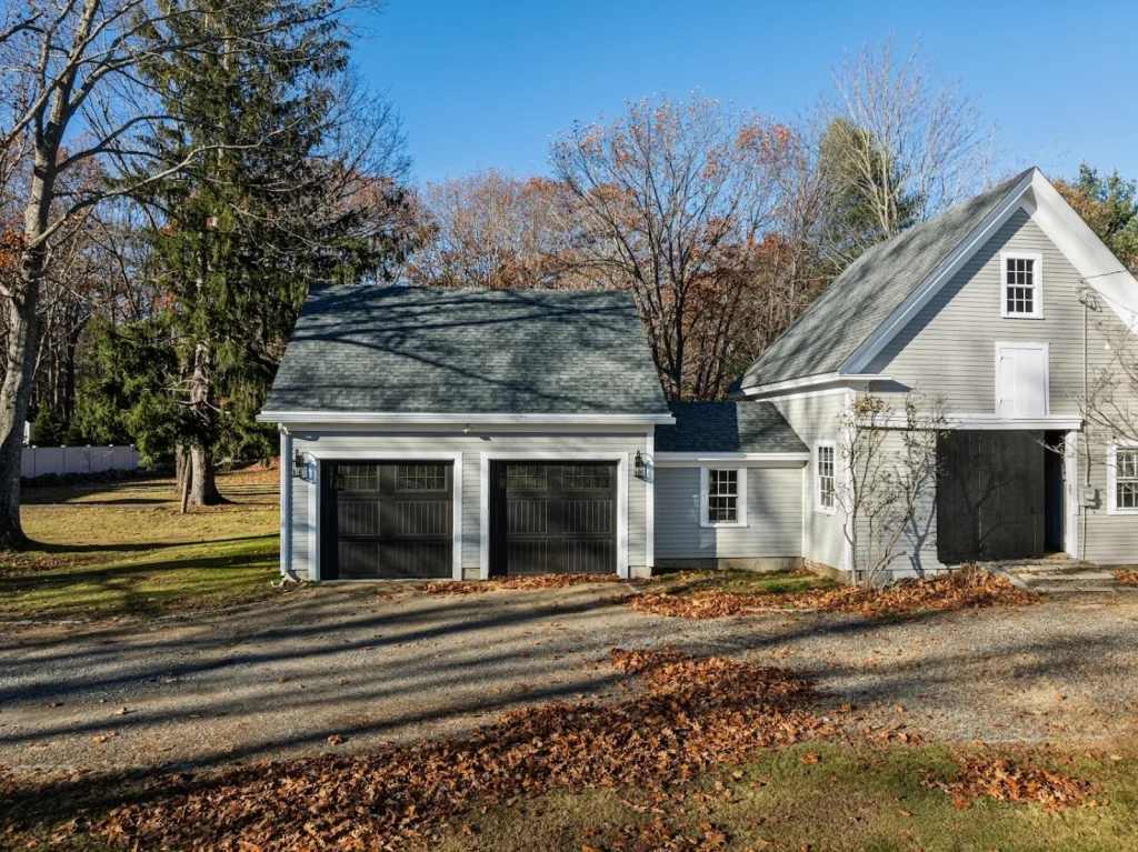 Grey home with two black garage doors, attached barn, and a gravel driveway with autumn leaves