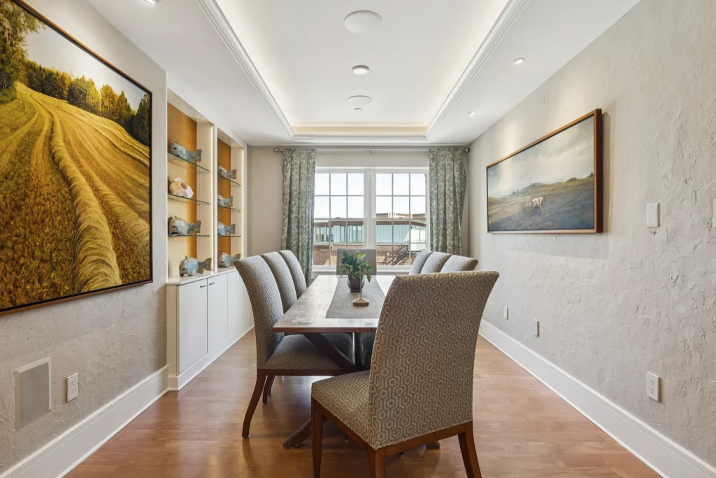 A modern dining room with a long wooden table, beige patterned chairs, and large landscape paintings on the walls, flanked by windowed doors at the far end and built-in shelves on the left.
