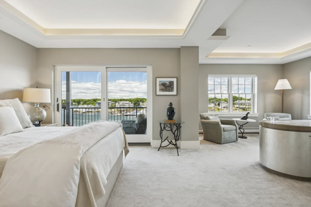 Bright hotel bedroom with sliding balcony doors opening to a waterfront view and neutral furnishings with a seating area nearby.
