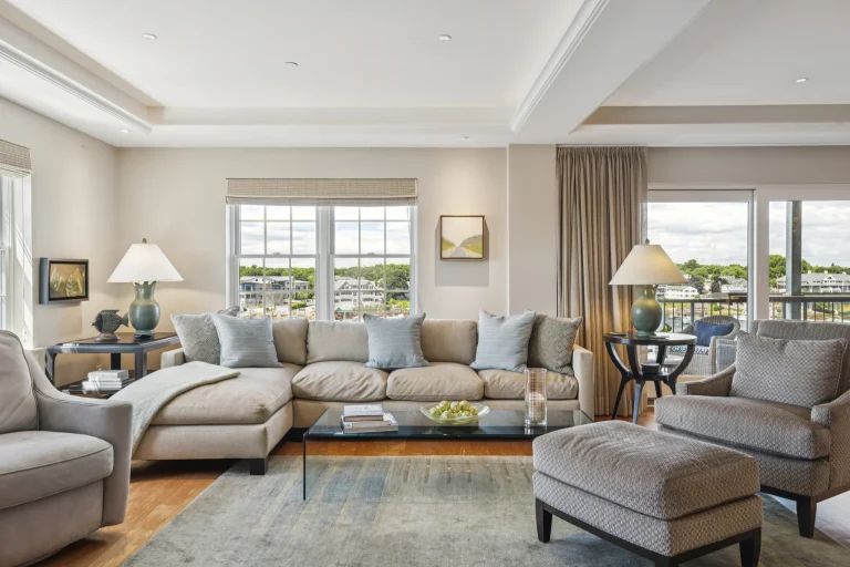 Cozy living room with a beige sectional, light blue pillows, glass coffee table, and large windows showing a neighborhood view.