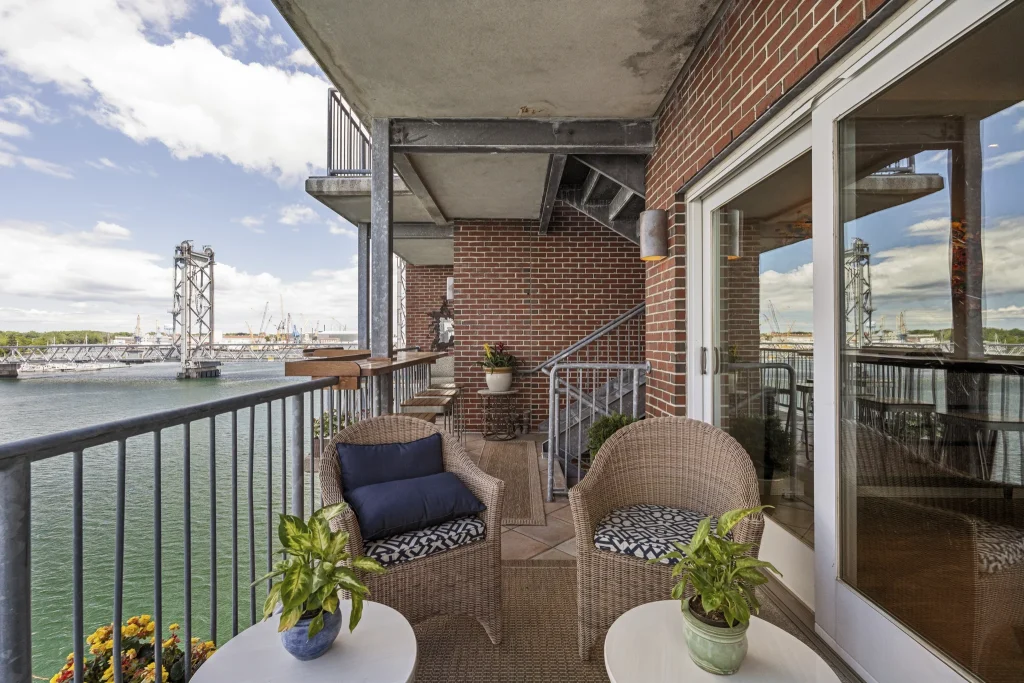 Balcony seating area with wicker chairs, small tables, and potted plants along a brick wall, overlooking a harbor and bridge.